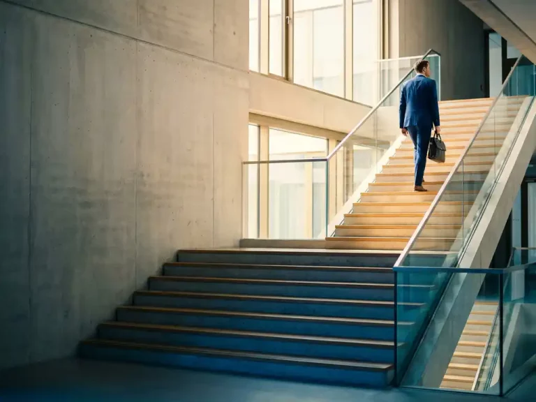 Geschäftsmann mit Aktentasche steigt moderne Treppe in Berliner Bürogebäude hinauf, goldenes Licht von oben.